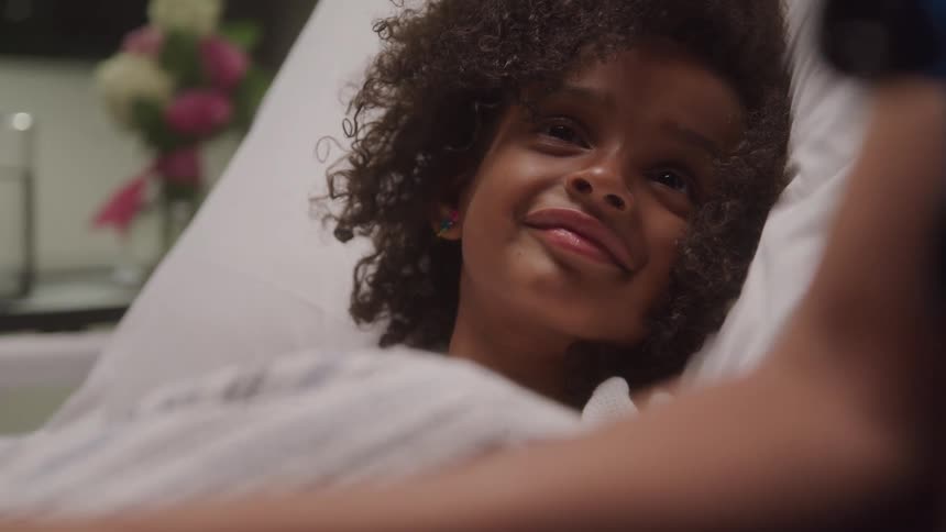 A young girl with dark curly hair lies in a bed looking up with a slight smile with a vase of flowers blurred in the background