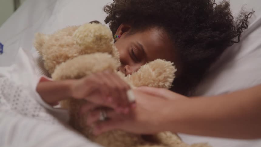 A child in bed hugs a teddy bear receiving it from a caregivers hands