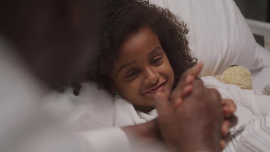 A young girl with dark curly hair lies in a bed looking up with a soft smile while holding hands with an adult