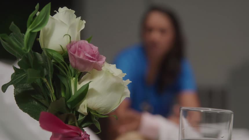A small bouquet of white and pink roses with a red ribbon is in the foreground with a glass of water and a blurred figure of a woman in blue in the background