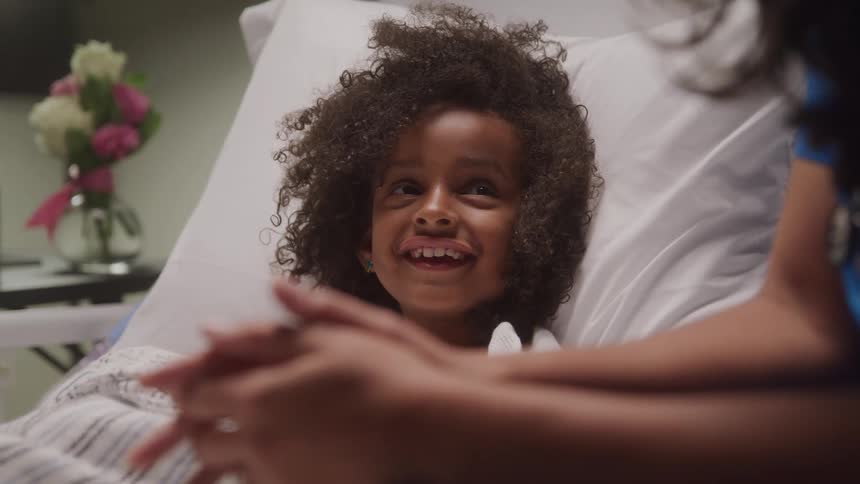 A joyful child with curly hair is smiling brightly while sitting up in a hospital bed with a persons hands in the foreground and a bouquet of flowers visible in the background