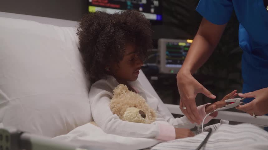 A child with dark curly hair is seen in a hospital bed holding a teddy bear a person in blue scrubs is adjusting a medical device on the childs finger