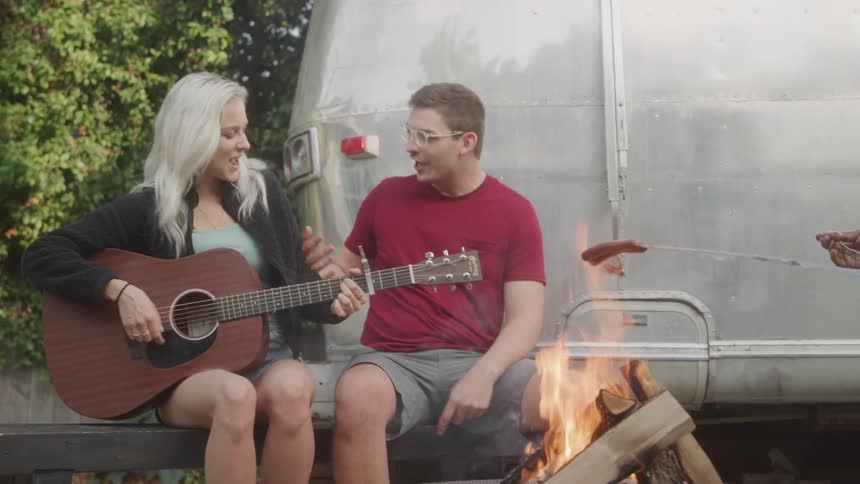The video features two friends enjoying a campfire outdoors with one playing a guitar and the other holding a hot dog on a stick over the flames they are seated in front of a shiny metallic travel trailer under clear skies
