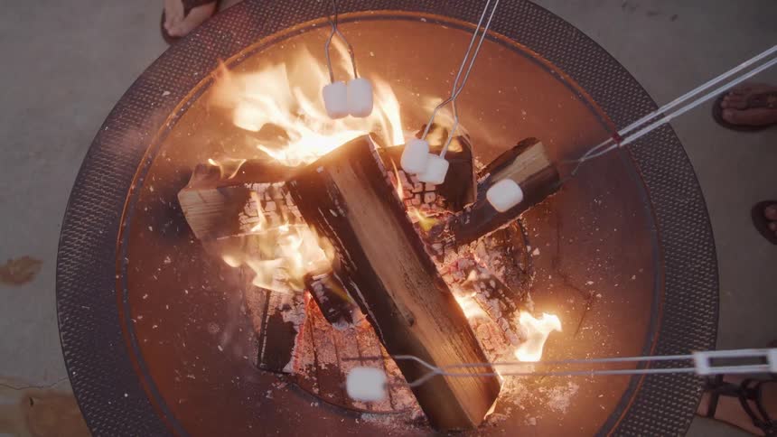 Several marshmallows being roasted on skewers over a crackling fire in a round metal fire pit with logs burning brightly