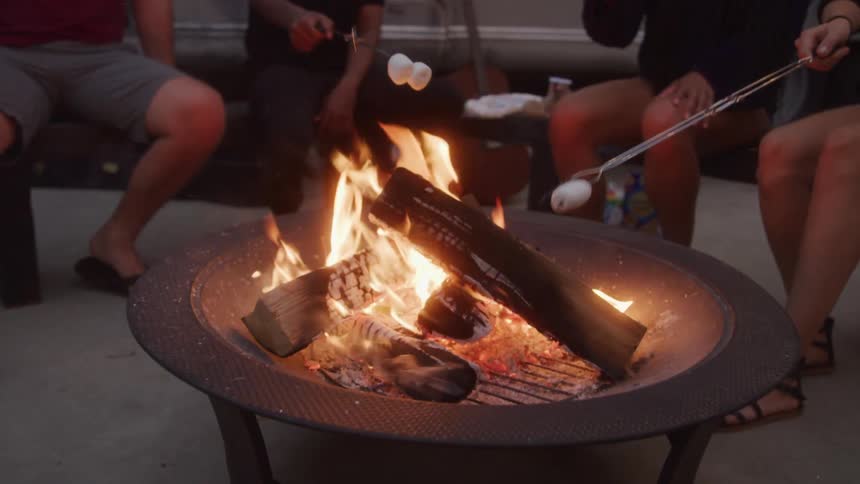 A close view shows several peoples legs and hands as they roast marshmallows over a crackling fire in a metal fire pit
