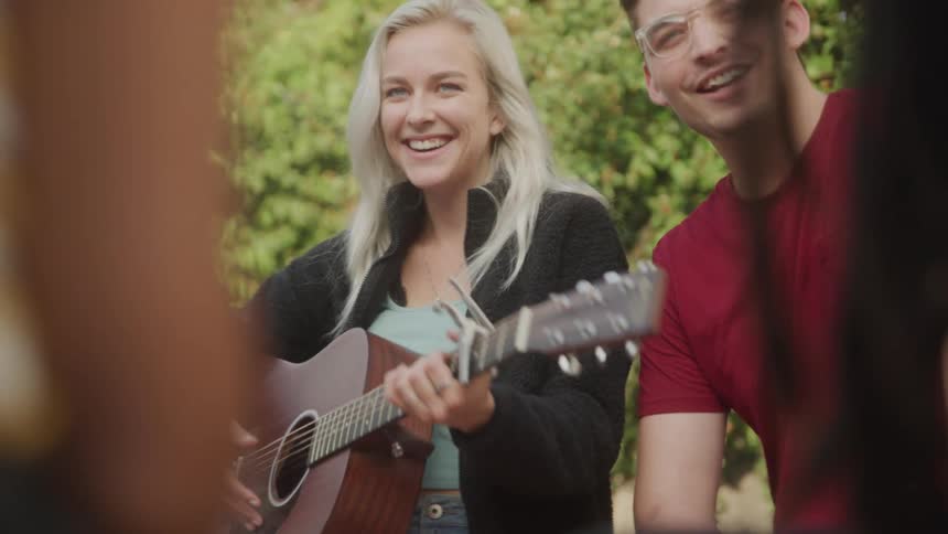 Friends sit by campfire as woman plays guitar