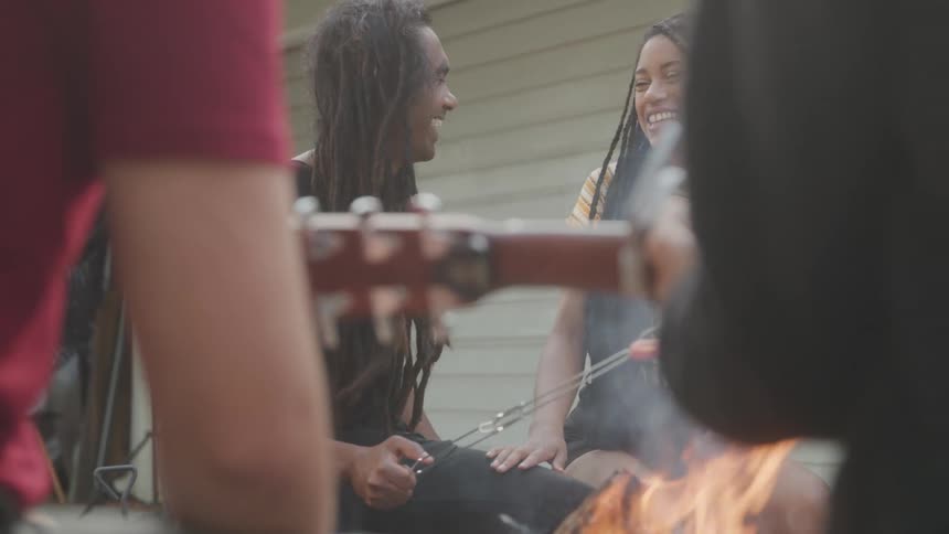 The video features a group of people enjoying themselves around a campfire one person is playing a guitar while others are laughing and roasting food over the open flames