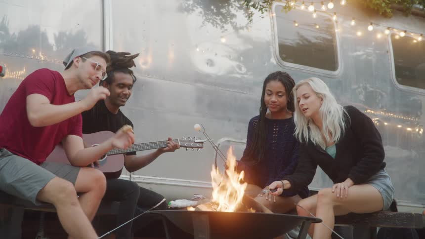 Friends chat by campfire near trailer as one plays guitar
