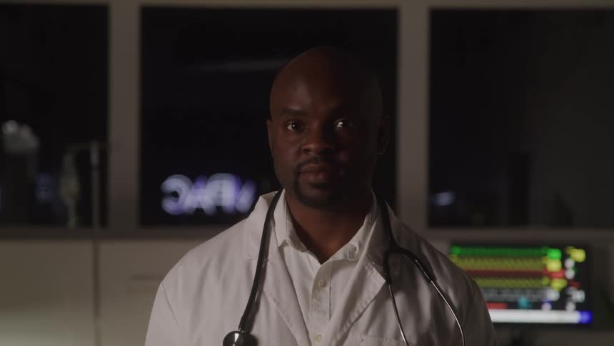 A male doctor stands in a dimly lit hospital or medical setting