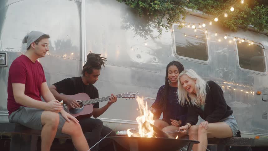 A group of friends sit around a glowing fire pit outside a silver airstream trailer one strumming a guitar while the others chat and relax beneath hanging string lights that cast a festive glow