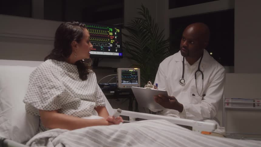 A female patient is seen sitting up in a hospital bed engaged in conversation with a male doctor who is holding a clipboard medical monitoring equipment is visible in the background displaying vital signs on screens