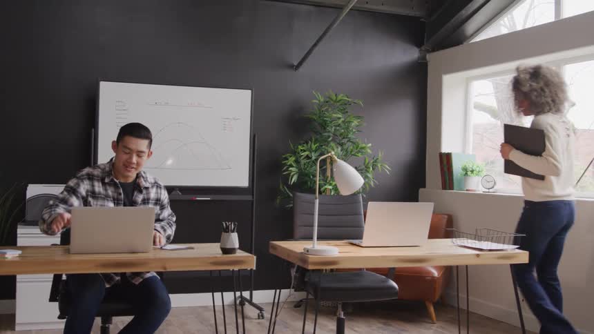 A young adult man works at a desk with a laptop as a young adult woman walks past and sits in another desk with a laptop