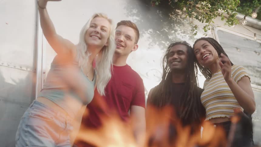 The video features a group of diverse young adults gathered outdoors possibly around a campfire smiling and interacting one person is actively taking a selfie with a phone capturing the moment