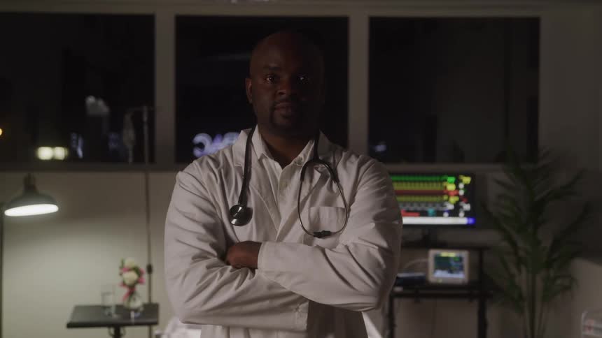 A confident male doctor with folded arms stands prominently in a dimly lit medical room with a vital signs monitor and an outdoor night view visible in the background