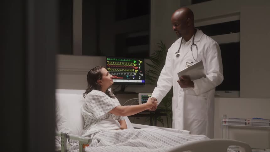 A female patient is seen sitting up in a hospital bed engaged in conversation with a male doctor who is holding a clipboard medical monitoring equipment is visible in the background displaying vital signs on screens