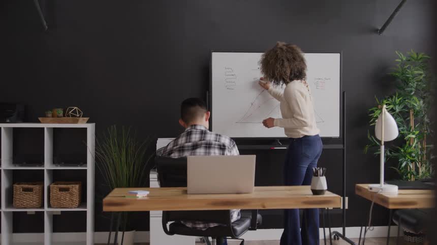 A woman with curly hair is actively writing on a whiteboard while a man sits at a desk with a laptop