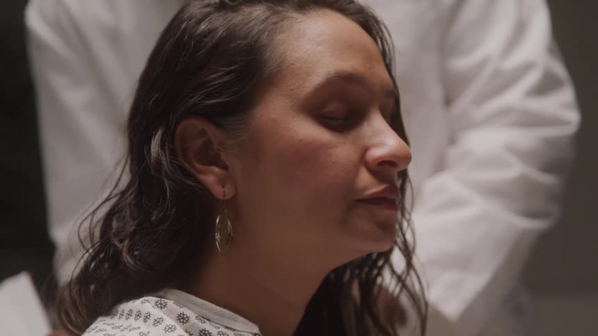 A woman with wet hair and closed eyes sits calmly in a white medical gown as someone in a lab coat stands behind her
