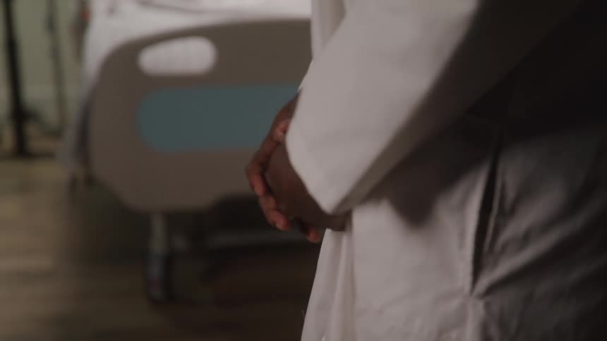 A doctor walks beside a hospital bed with hands clasped likely checking on a patient