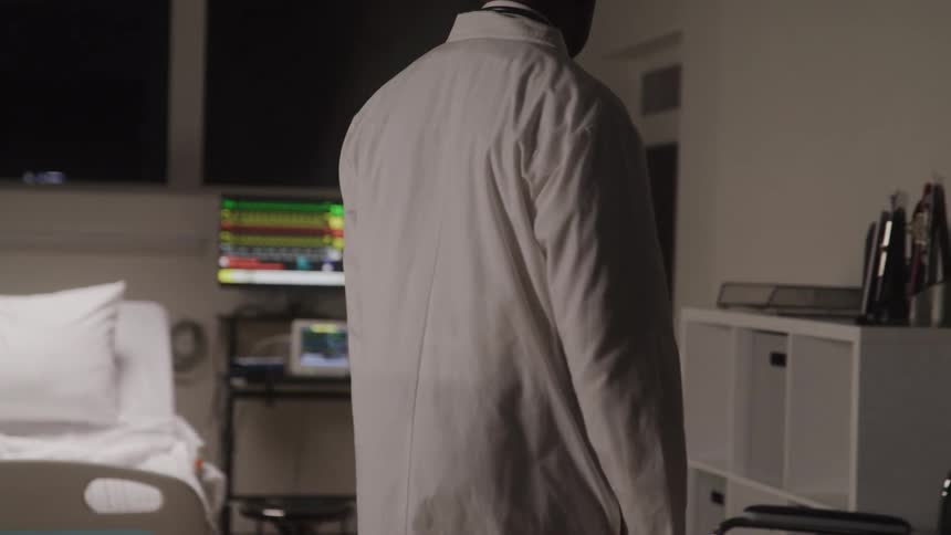 A doctor walks into a dimly lit hospital room looking pensive