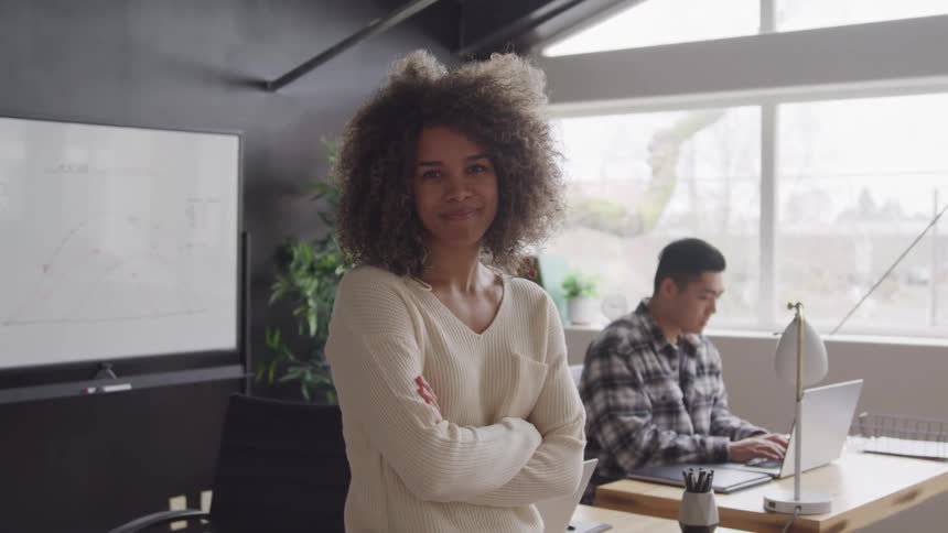 A confident woman with large curly hair stands with her arms crossed looking directly at the camera in a modern office while a man works on a laptop