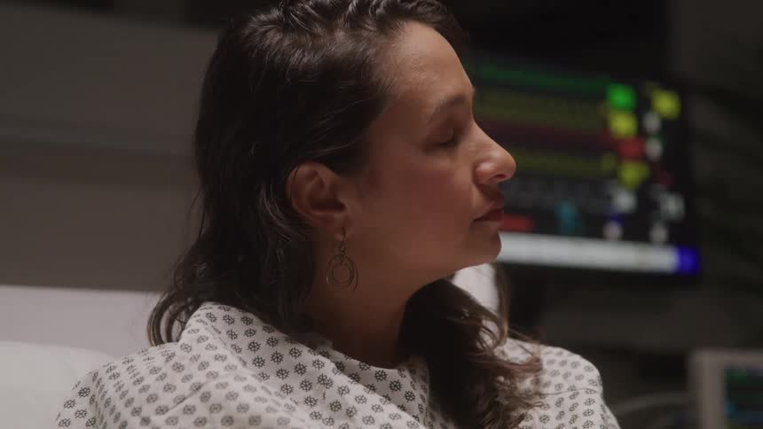 A woman lies in a hospital bed looking to the side in a clinical setting