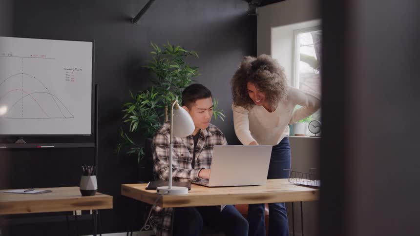 A woman stands by a man working on a laptop in a modern office