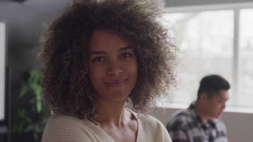 A woman smiles warmly while standing indoors with someone seated behind her