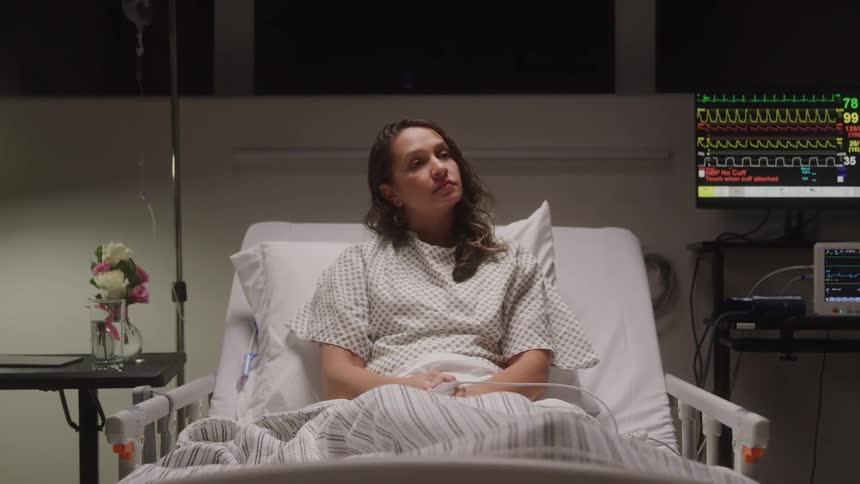 A woman sits thoughtfully in a dimly lit hospital bed