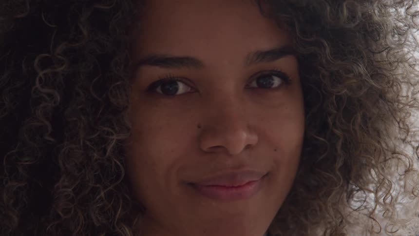 A woman with curly hair is shown in a closeup looking toward the side and then directly at the camera with a gentle smile the lighting suggests an indoor setting possibly near a window giving her face a soft glow