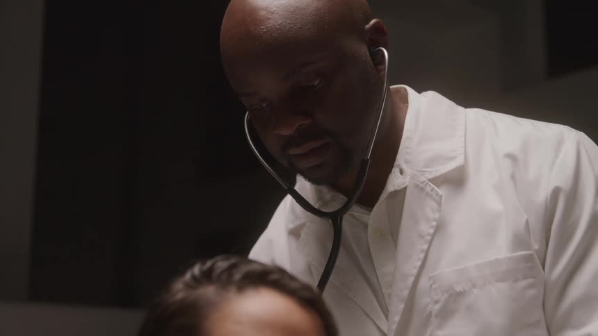 The video shows a closeup shot of a bald man wearing a white lab coat and a stethoscope around his neck he appears to be looking down at someone or something out of frame seemingly concentrating