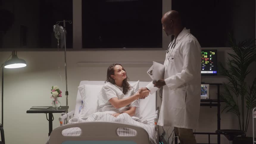 A male doctor in a white lab coat is standing next to a female patient who is sitting up in a hospital bed they shake hands while medical equipment with glowing screens is visible in the background