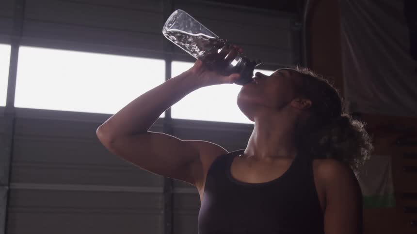Woman with curly hair wearing a dark tank top drinking from a clear water bottle in a gym with bright windows in the background
