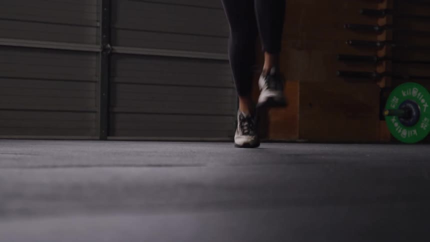 A person with curly hair is shown in a low angle bent over in a starting position suggesting they are about to begin an exercise or sprint in the background theres gym equipment including a green weight plate indicating an indoor fitness setting