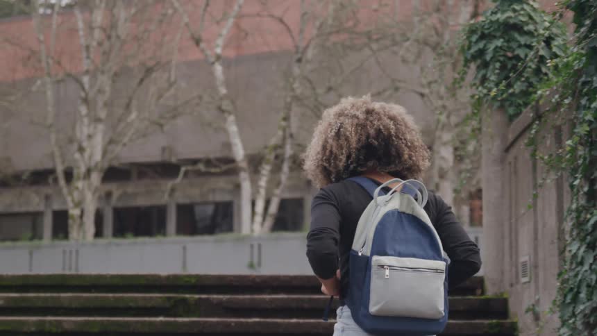 A person with curly hair and a backpack walks up a flight of outdoor stairs the stairs are made of concrete and have some moss growing on them
