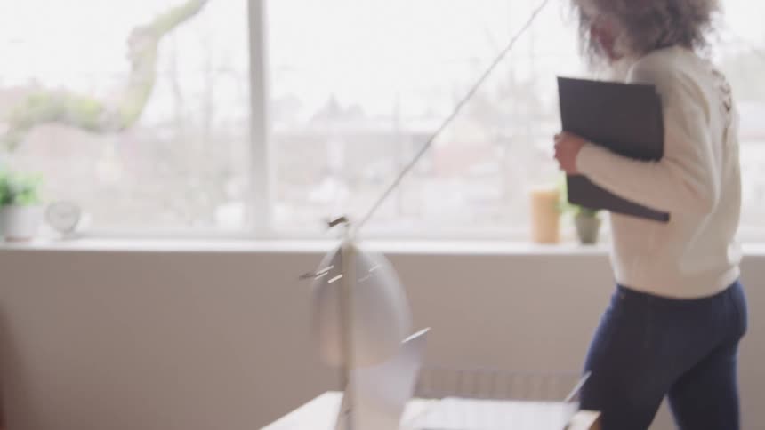 A woman with curly hair walks into a bright modern office space carrying a tablet she approaches a desk where a man is working on a laptop and they begin to collaborate