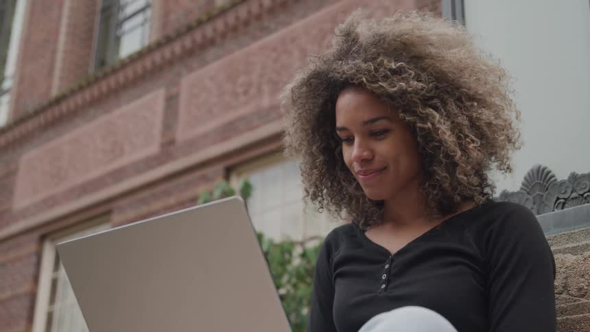 A woman with curly brown hair smiles while looking at a laptop computer with a pink brick building in the background