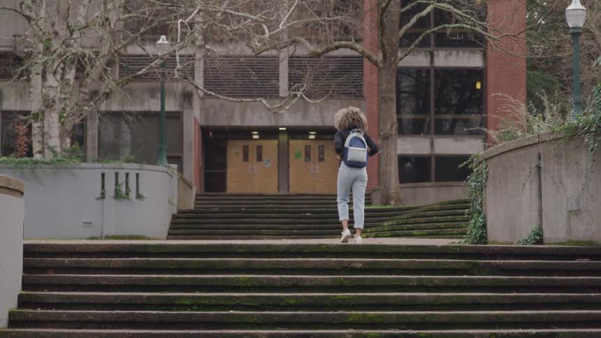 A teenage girl walks into a school building at midday carrying a backpack and wearing her long curly hair down