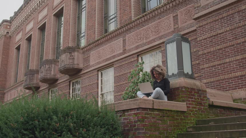 Female sitting on ledge near steps of a brick building working on her laptop