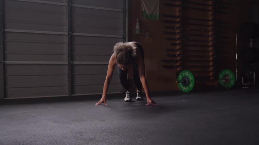 A woman with curly light colored hair is running during a workout wearing a dark sports bra and black leggings with gym equipment in the background