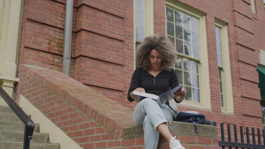Female sitting on brick ledge near steps in front of a building reading a textbook