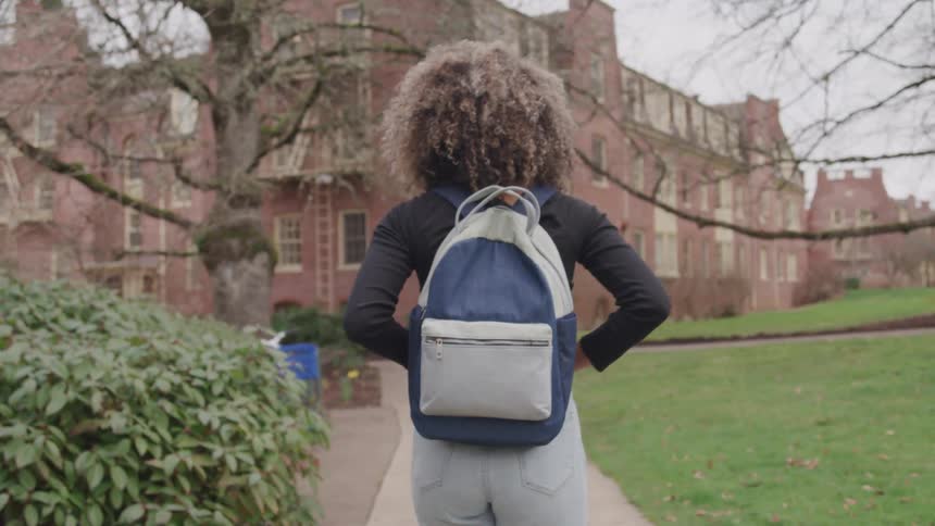A young woman carrying a backpack walking toward a school building surrounded by green vegetation at midday