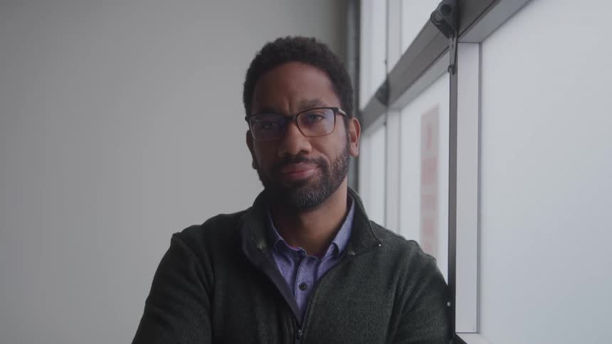 A man with a beard and glasses is looking directly at the camera standing beside a window or a white wall he is wearing a dark zipped jacket over a collared shirt
