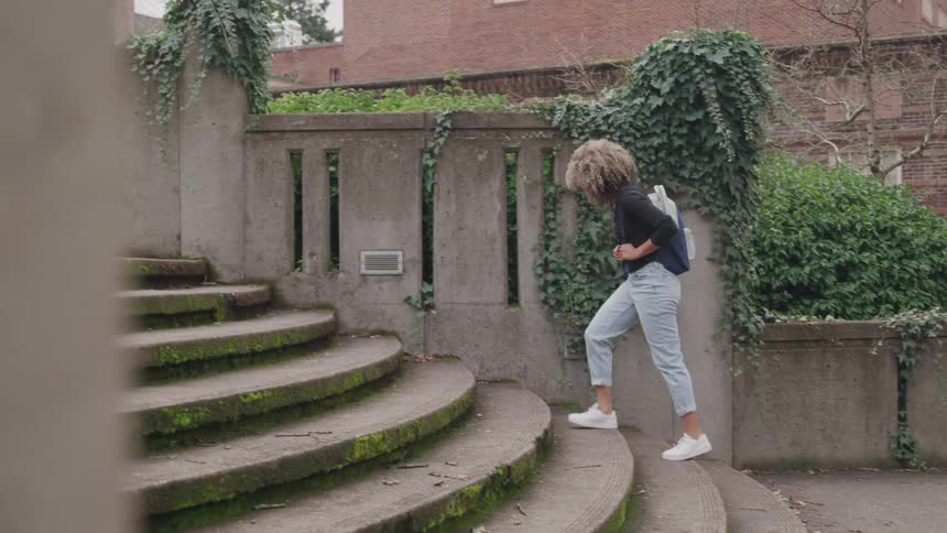 A young woman with a backpack climbs a curved mossy stone staircase surrounded by ivycovered walls in a serene oldworld setting