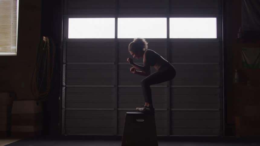 The video shows a person with curly hair performing box jumps in a dimly lit gym or warehouse space they are silhouetted against bright windows or garage doors in the background