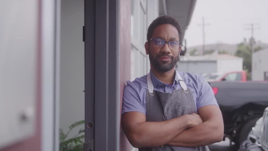 The video features a man with a beard and glasses wearing a blue shirt and a grey apron standing with his arms crossed he is leaning against a doorway with a blurred background showing a street and buildings suggesting he might be a business owner
