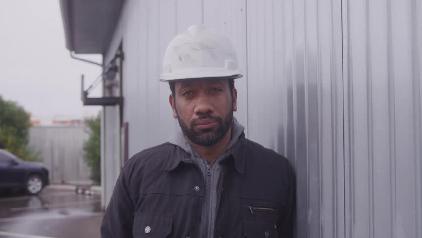 A man with a beard and a white hard hat leans against a corrugated metal wall looking directly at the viewer with a neutral expression under an overcast sky