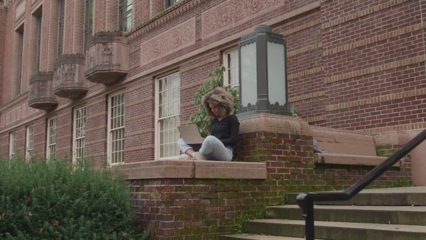 Female sitting on a ledge near steps front of a brick building working on her laptop