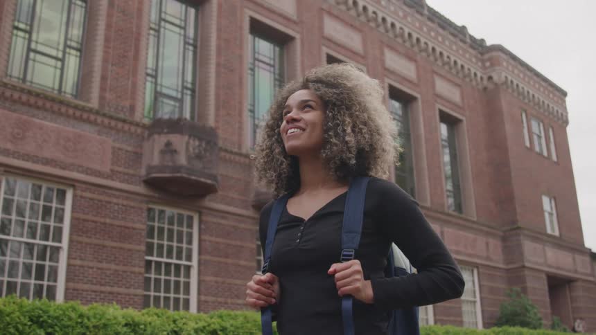 A young woman with long curly hair standing in the middle of the street at midday smiling and holding the strap of her backpack