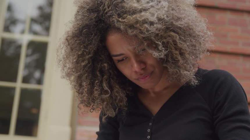 A woman with voluminous curly hair is shown from the chest up looking down with a serious or concerned expression she is wearing a black collared shirt with buttons