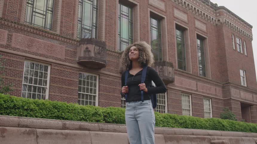 A young female student looks around while standing on a walkway in a campus during daytime